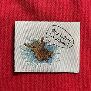 Könnte beinhalten: Ein weißer Stoff-Patch mit einem braunen Bären, der in blauem Wasser schwimmt. Der Patch hat eine Sprechblase mit dem Text "Das Leben ist schön!" in Schwarz.
