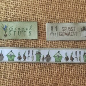 May include: Two green fabric labels with white text and illustrations. The first label says "Einzel St&uuml;ck" and has a drawing of a flower bulb. The second label says "Selbst Gemacht?" and has a drawing of a flower pot. A green and white ribbon with a repeating pattern of flower bulbs, pots, and gardening tools.