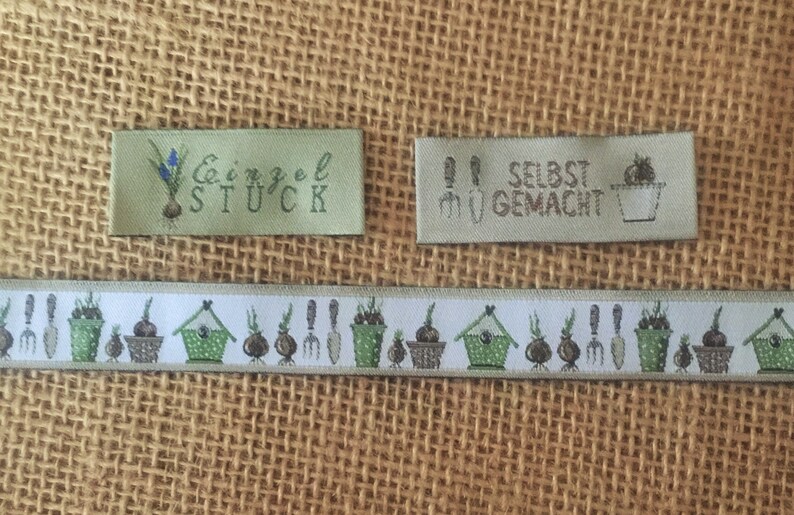 May include: Two green fabric labels with white text and illustrations. The first label says "Einzel St&uuml;ck" and has a drawing of a flower bulb. The second label says "Selbst Gemacht?" and has a drawing of a flower pot. A green and white ribbon with a repeating pattern of flower bulbs, pots, and gardening tools.