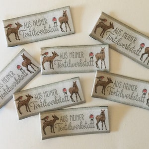 May include: A set of eight beige fabric labels with a brown deer and a red bird design. The labels have the text "AUS MEINER Textilwerkstatt" printed on them.