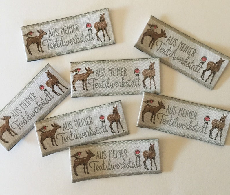 May include: A set of eight beige fabric labels with a brown deer and a red bird design. The labels have the text "AUS MEINER Textilwerkstatt" printed on them.