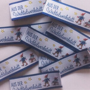Könnte beinhalten: Eine Sammlung von blauen und weißen Stoff-Geschenk-Etiketten mit dem Text "AUS DER WICHTELWERKSTATT" und einer kleinen Illustration eines Zwergs. Die Etiketten sind mit goldenen Sternen verziert.