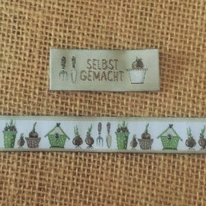 May include: A white fabric ribbon with a repeating pattern of green potted plants, brown flower pots, a green birdhouse, and gardening tools. A green label with the text "SELBST GEMACHT" is above the ribbon.