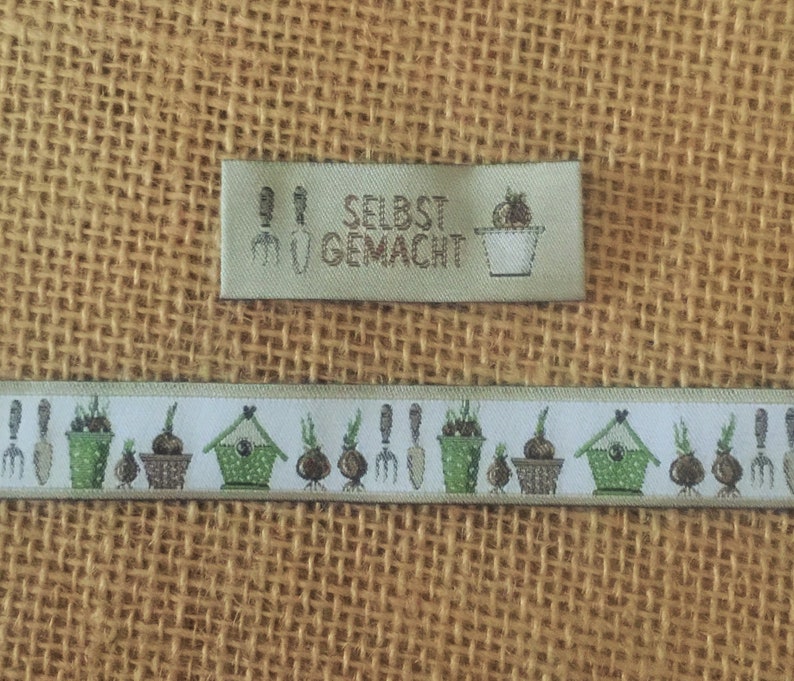 May include: A white fabric ribbon with a repeating pattern of green potted plants, brown flower pots, a green birdhouse, and gardening tools. A green label with the text "SELBST GEMACHT" is above the ribbon.