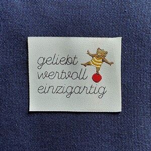 Könnte beinhalten: Weißes Stofflabel mit dem Text "geliebt wertvoll einzigartig" in schwarzer kursivschrift. Eine Cartoon-Illustration eines braunen Bären, der auf einem roten Ball balanciert, befindet sich über dem Text.