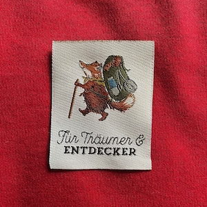 Könnte beinhalten: Ein weißer Stoff-Patch mit einem braunen Fuchs, der einen grünen Rucksack und einen Wanderstock trägt. Der Text "Für Träumer & ENTDECKER" ist in Schwarz unter dem Fuchs aufgedruckt.