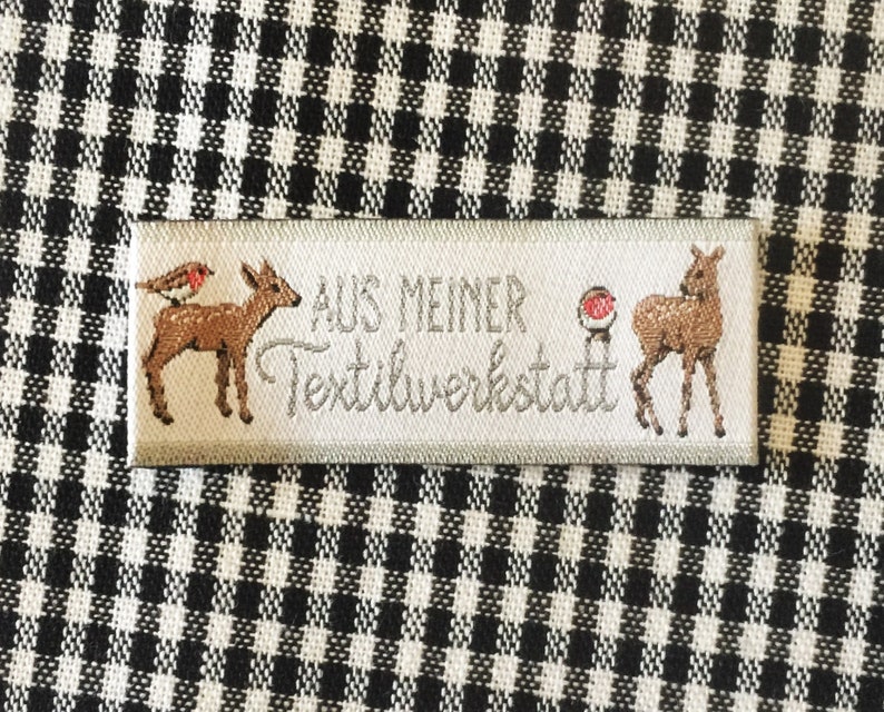 May include: A white fabric label with the text "AUS MEINER Textilwerkstatt" in gray lettering. The label features two brown deer, one with a red robin perched on its back, and a red circle with a white dot in the center.