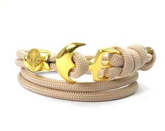 May include: A beige paracord bracelet with a gold anchor charm and a gold circular charm with the letters "HC" and a design inside.