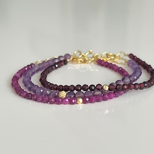 Puede incluir: Tres pulseras superpuestas con cuentas moradas, rosadas y moradas oscuras. Las pulseras están conectadas con cierres dorados y tienen pequeños detalles dorados.