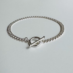 Könnte beinhalten: Ein silbernes Panzerkettenarmband mit Knebelverschluss. Das Armband besteht aus einer Reihe ineinandergreifender Glieder, die eine strukturierte Oberfläche bilden. Der Knebelverschluss ist ein schlichtes, aber elegantes Design, auf weißem Hintergrund.