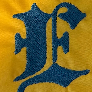 May include: A bright yellow fabric surface featuring a large, dark blue embroidered letter E in an Old English gothic font. The embroidery shows a dense, textured stitch pattern that stands out against the smooth, solid yellow background material.