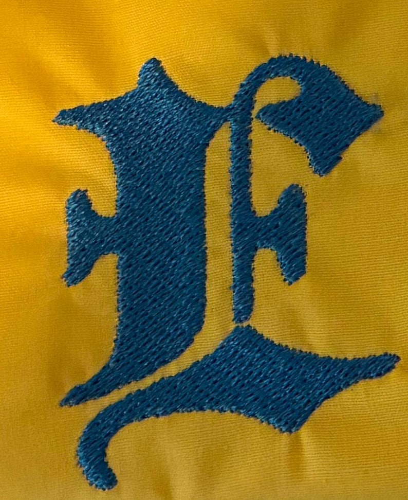 May include: A bright yellow fabric surface featuring a large, dark blue embroidered letter E in an Old English gothic font. The embroidery shows a dense, textured stitch pattern that stands out against the smooth, solid yellow background material.