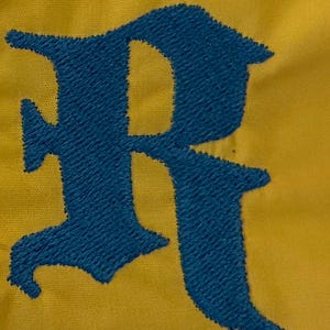 May include: A mustard yellow fabric surface featuring a large, dark blue embroidered letter R in a gothic-style font. The embroidery shows a dense, textured thread pattern that stands out against the smooth, solid-colored textile background.