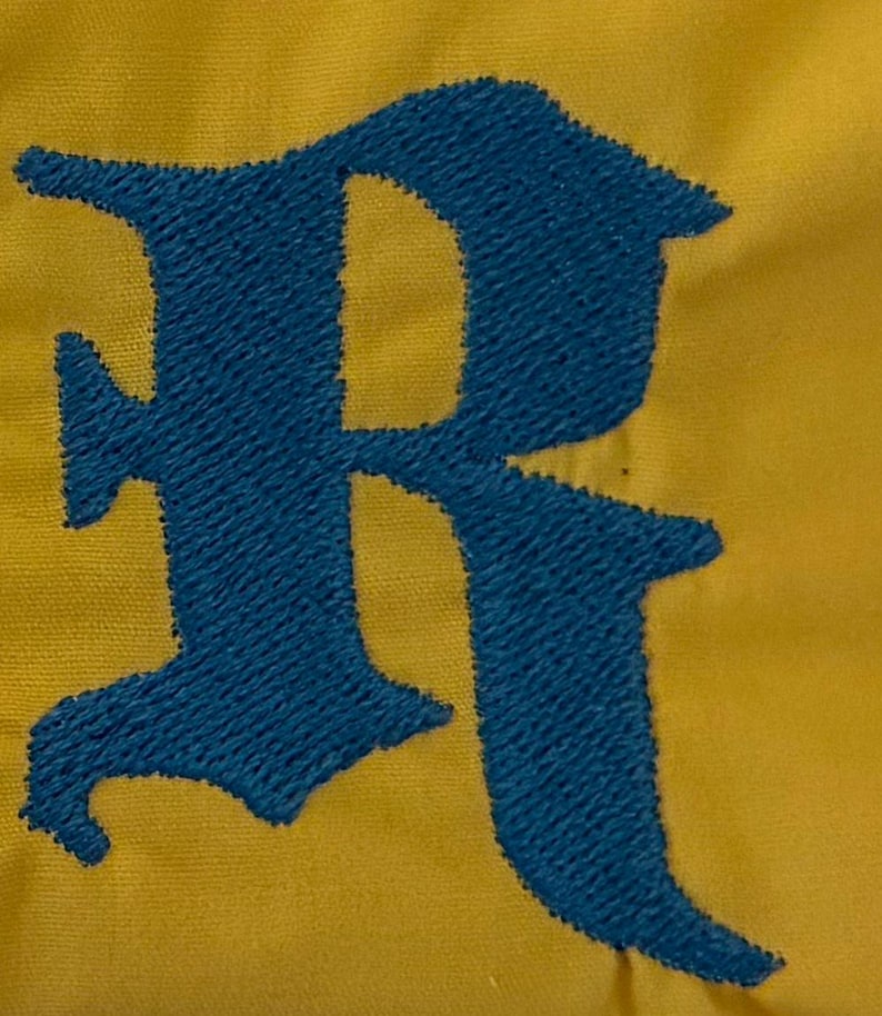 May include: A mustard yellow fabric surface featuring a large, dark blue embroidered letter R in a gothic-style font. The embroidery shows a dense, textured thread pattern that stands out against the smooth, solid-colored textile background.