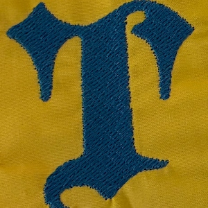 May include: A dark blue embroidered letter T in an old English gothic font style, stitched onto a solid mustard yellow fabric background. The embroidery features a dense, textured thread pattern that stands out against the smooth textile surface.