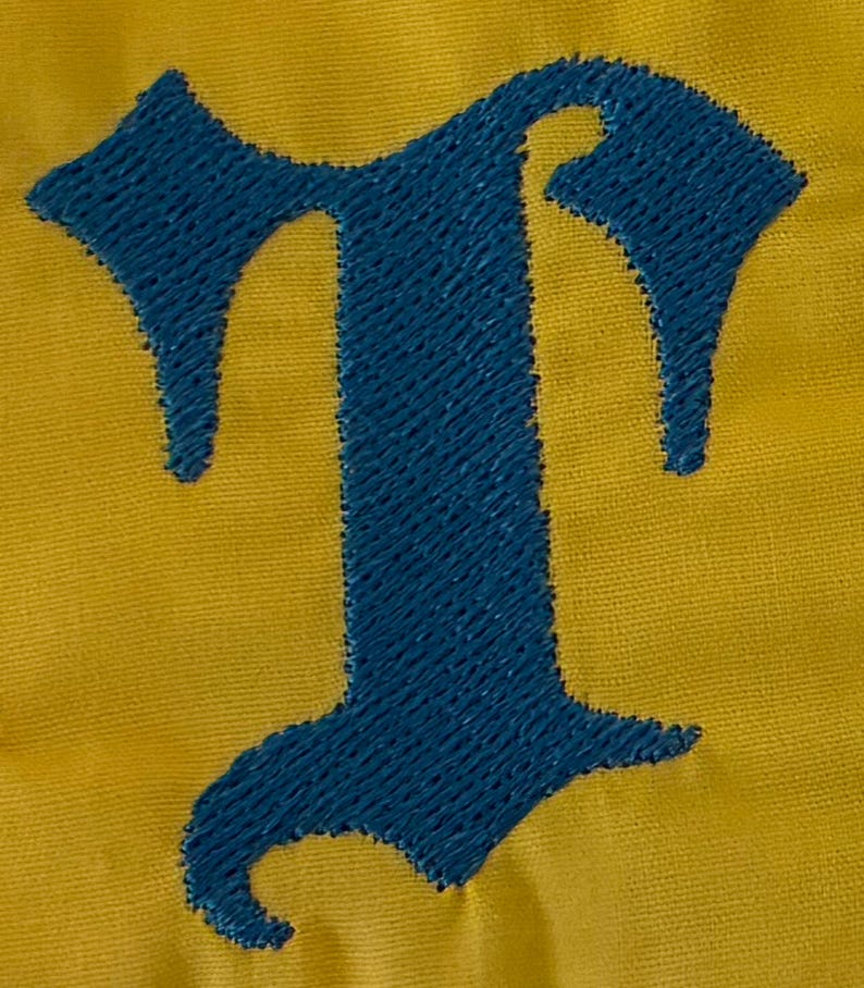 May include: A dark blue embroidered letter T in an old English gothic font style, stitched onto a solid mustard yellow fabric background. The embroidery features a dense, textured thread pattern that stands out against the smooth textile surface.