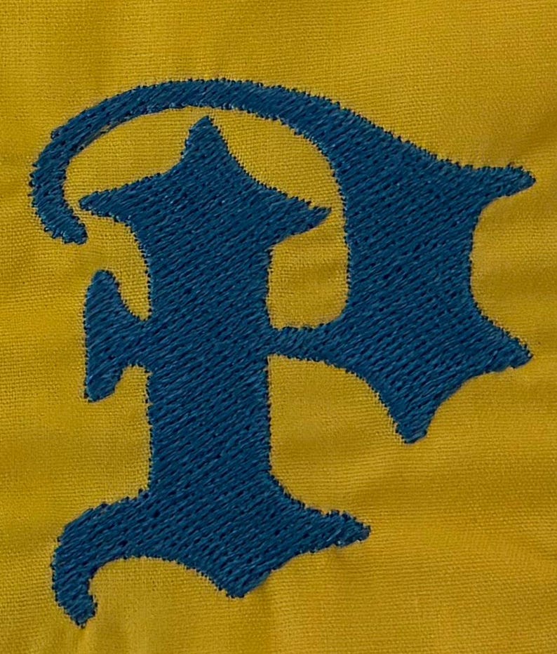 May include: A mustard yellow fabric background featuring a large, dark blue embroidered letter P in a gothic, old English style font. The embroidery has a visible, textured stitch pattern that stands out against the smooth yellow textile.