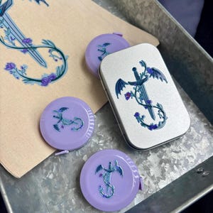 May include: A collection of fantasy-themed items, including two purple tape measures, a silver tin, and a beige pouch. Each item features a design of a sword with dragon wings and floral accents. The items are arranged in a metal tray.