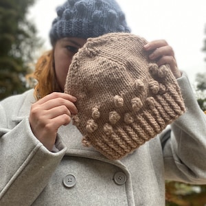 Könnte beinhalten: Zwei Strickmützen, eine braune mit einem strukturierten Noppenmuster und eine blaue mit einem ähnlichen Muster. Die Mützen werden vor einer Person gehalten, die einen grauen Mantel trägt.