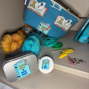 May include: A teal zippered pouch with a gold zipper, featuring a book illustration and the text "Just One More Chapter." Also shown are yarn, a tin with "Craft More Read More" text, a tape measure, scissors, and stitch markers.