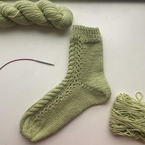 May include: A green knitted sock with a cable pattern. The sock is lying on a white surface with a ball of green yarn and a knitting needle.