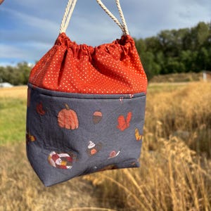 May include: A drawstring bag with a red and white polka dot fabric top and a gray bottom with a fall-themed print of pumpkins, acorns, and other autumnal elements.