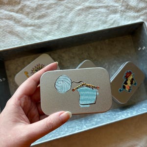 May include: A silver metal tin with a blue and white illustration of a ball of yarn and knitting needles. The tin is part of a set of four tins with different illustrations.