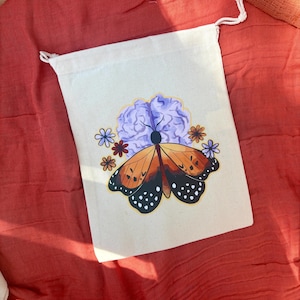 May include: A cream-colored drawstring bag featuring a butterfly design with orange wings and black spots, surrounded by a purple flower and smaller flowers. The bag is made of a canvas-like material.