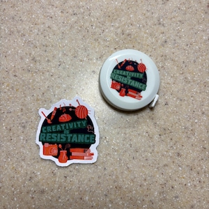 May include: A white tape measure and a sticker with the text "CREATIVITY IS RESISTANCE". The sticker and tape measure feature illustrations of art supplies in red, orange, and black. The tape measure is round and the sticker is die-cut.