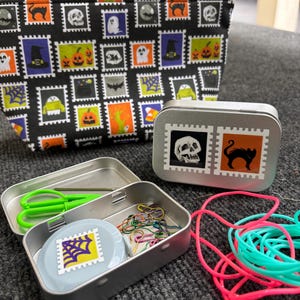 May include: A black Halloween-themed zippered pouch and two small metal tins. The pouch and one tin feature a repeating stamp design with ghosts, skulls, and pumpkins. The open tin contains scissors, paperclips, and a round container with a spiderweb stamp.