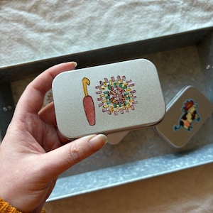 May include: Two small silver tins with colorful illustrations. One tin features a crochet hook and a colorful crocheted circle. The other tin features a cartoon cow.