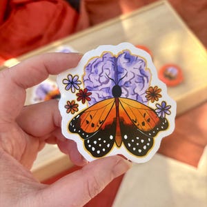 May include: A sticker featuring a monarch butterfly with orange and black wings, a purple brain, and colorful flowers. The sticker has a white border and is held in a hand.