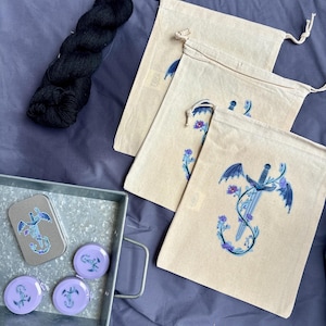 May include: A collection of craft supplies including three beige drawstring bags with a blue and purple dragon and sword design, a skein of black yarn, and a metal tray with matching pins and a tin.