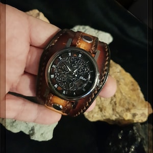 May include: A brown leather wristwatch with a black face and silver hands. The watch face has Roman numerals and a skeletonized design. The watch is on a brown leather band with a silver buckle.