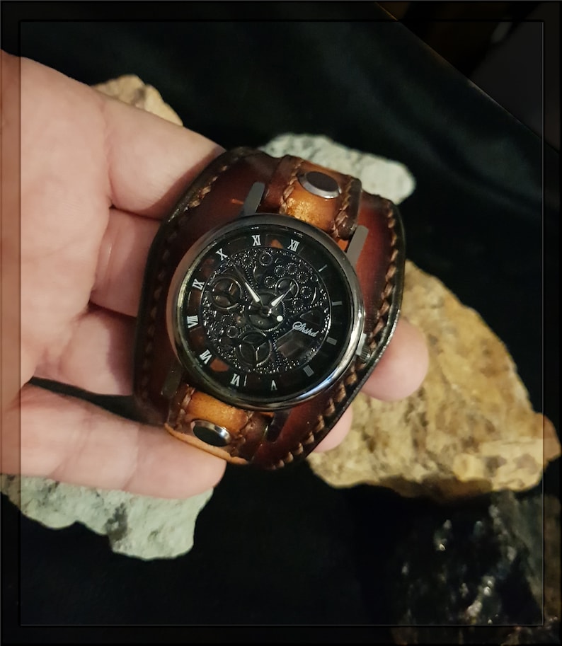 May include: A brown leather wristwatch with a black face and silver hands. The watch face has Roman numerals and a skeletonized design. The watch is on a brown leather band with a silver buckle.