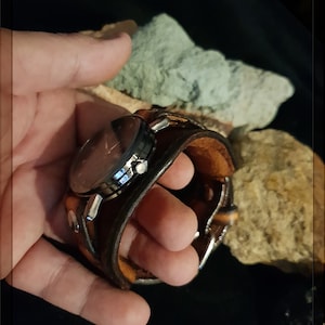 May include: A brown leather watch band with a silver watch face. The watch is held in a hand.