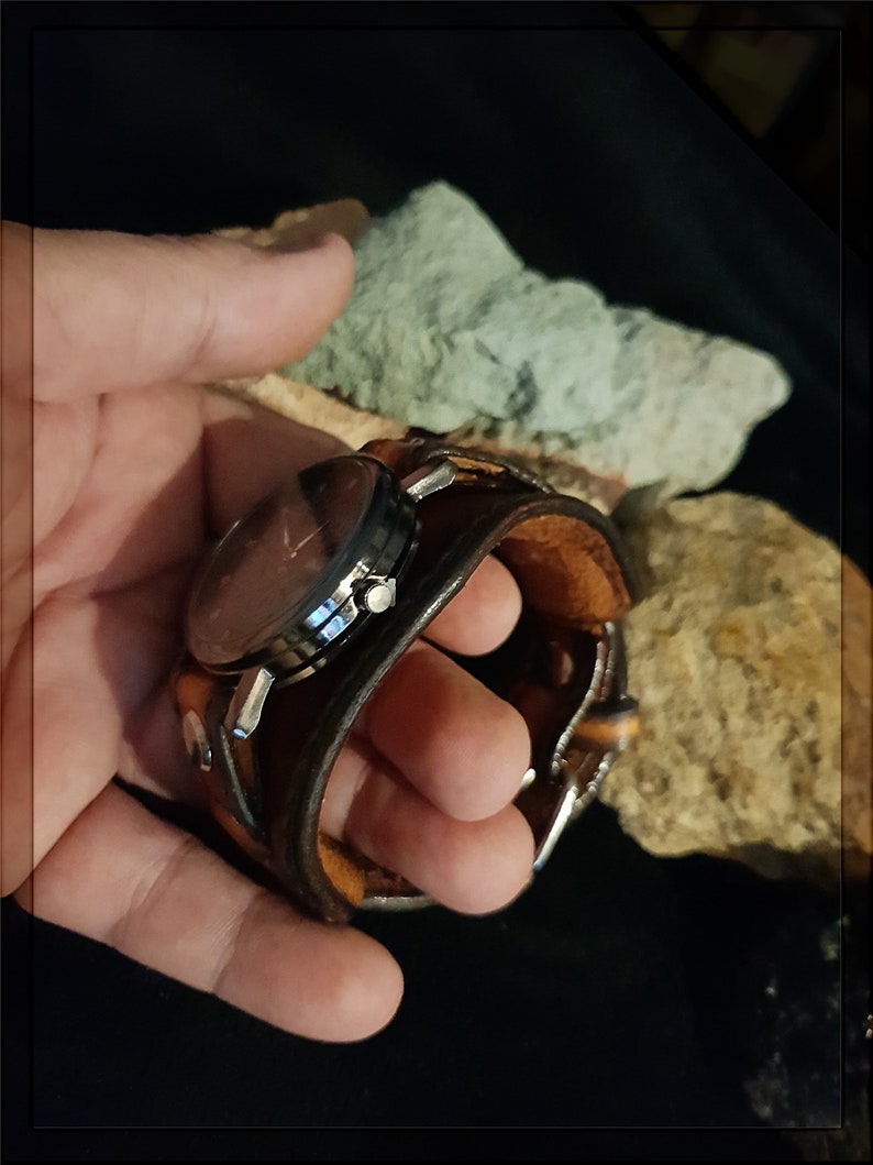 May include: A brown leather watch band with a silver watch face. The watch is held in a hand.
