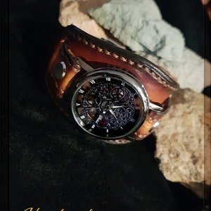 May include: A brown leather watch band with a silver watch face featuring a skeletonized movement. The watch face has Roman numeral hour markers and a black background. The watch is on a dark background with the word "Handmade" written in gold lettering.
