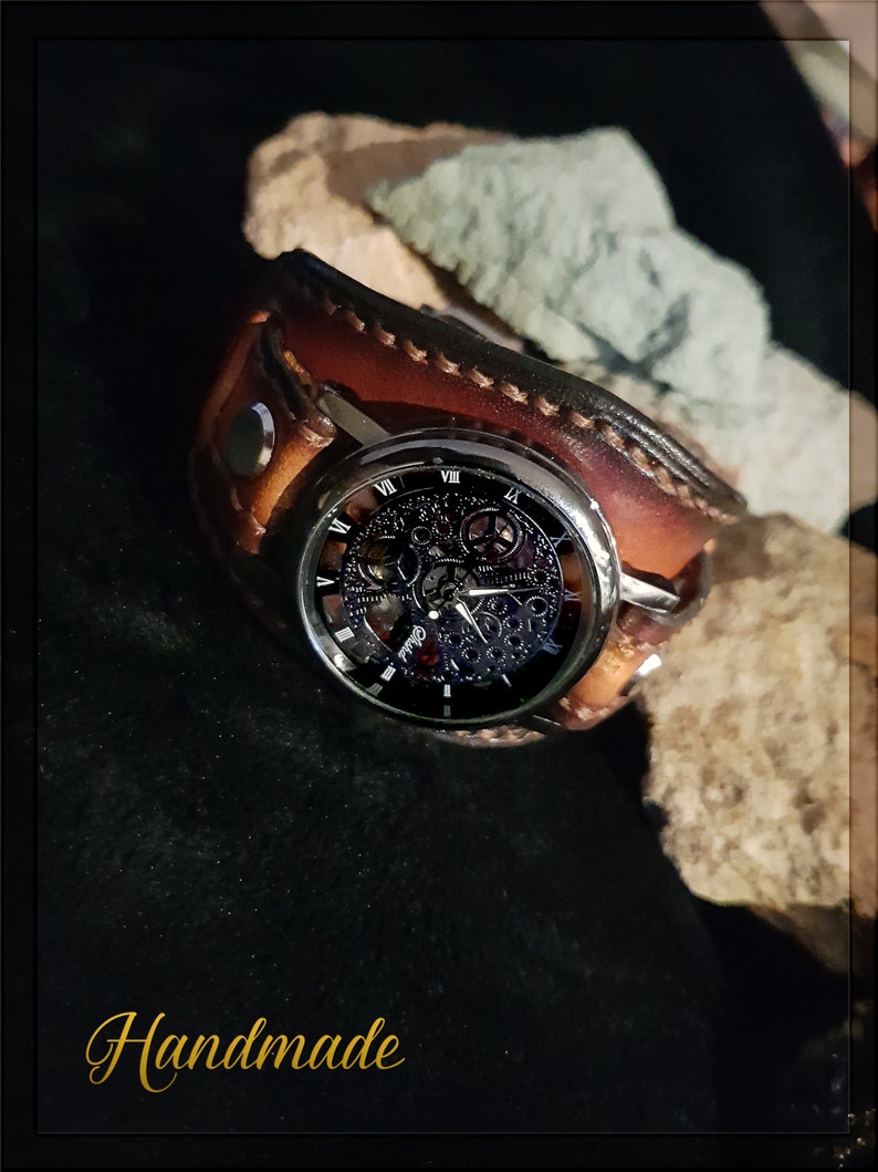 May include: A brown leather watch band with a silver watch face featuring a skeletonized movement. The watch face has Roman numeral hour markers and a black background. The watch is on a dark background with the word "Handmade" written in gold lettering.
