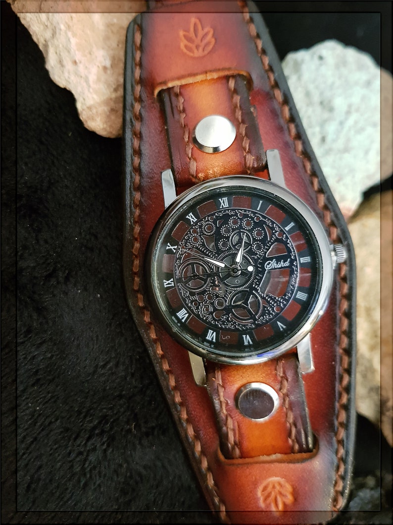 May include: A brown leather watch band with a silver watch face. The watch face has Roman numerals and a skeleton design. The watch band has a decorative stitching pattern.
