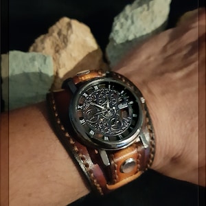 May include: A brown leather watch band with a silver watch face featuring a skeletonized movement. The watch face has Roman numeral hour markers and a black background.