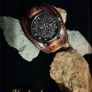 May include: A brown leather wristwatch with a black face and skeletonized hands. The watch has a silver metal band and a silver metal buckle. The watch is handmade and has the word "Handmade" written in gold lettering on the bottom of the image.