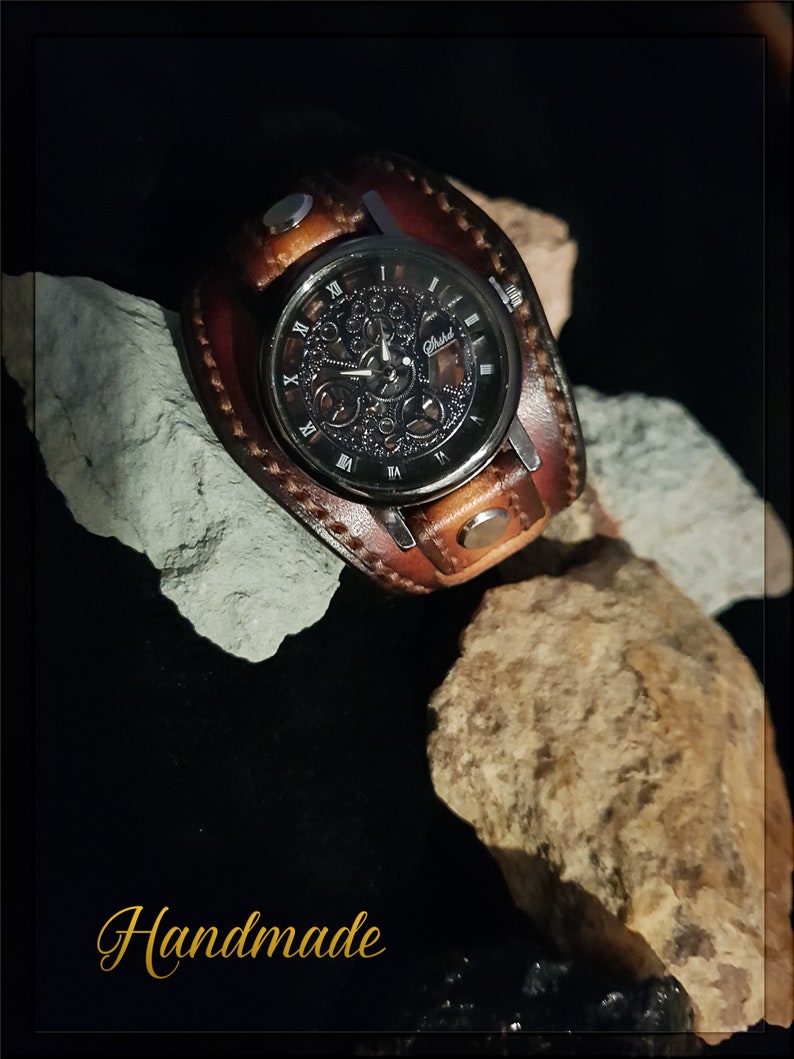 May include: A brown leather wristwatch with a black face and skeletonized hands. The watch has a silver metal band and a silver metal buckle. The watch is handmade and has the word "Handmade" written in gold lettering on the bottom of the image.