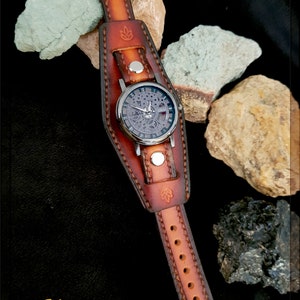 May include: A brown leather watch band with a silver watch face and a skeletonized movement. The band is handmade and has a decorative stitching pattern. The watch is on a black background with rocks.