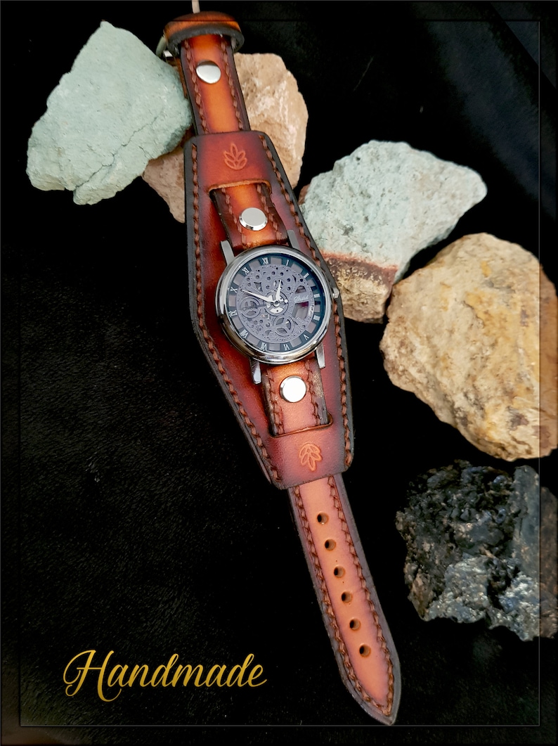 May include: A brown leather watch band with a silver watch face and a skeletonized movement. The band is handmade and has a decorative stitching pattern. The watch is on a black background with rocks.