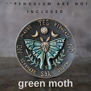 May include: A circular bronze-toned divination board featuring a central teal and gold moth, flanked by crescent moons and a dotted circle. The text reads YES, NO, MAYBE, and REPHRASE around the perimeter. Text at the top states PENDULUM ARE NOT INCLUDED.
