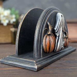May include: A black arched napkin holder with a distressed finish, featuring a raised relief design of a white ghost positioned between two orange pumpkins on the side panel, set on a rectangular base with matching dark weathered edges.