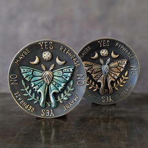 May include: Two circular dark metal plaques featuring a central moth relief, a moon, and stars. The text YES, NO, MAYBE, and REPHRASE is embossed around the perimeter. One moth is teal and gold, while the other is entirely gold-toned.