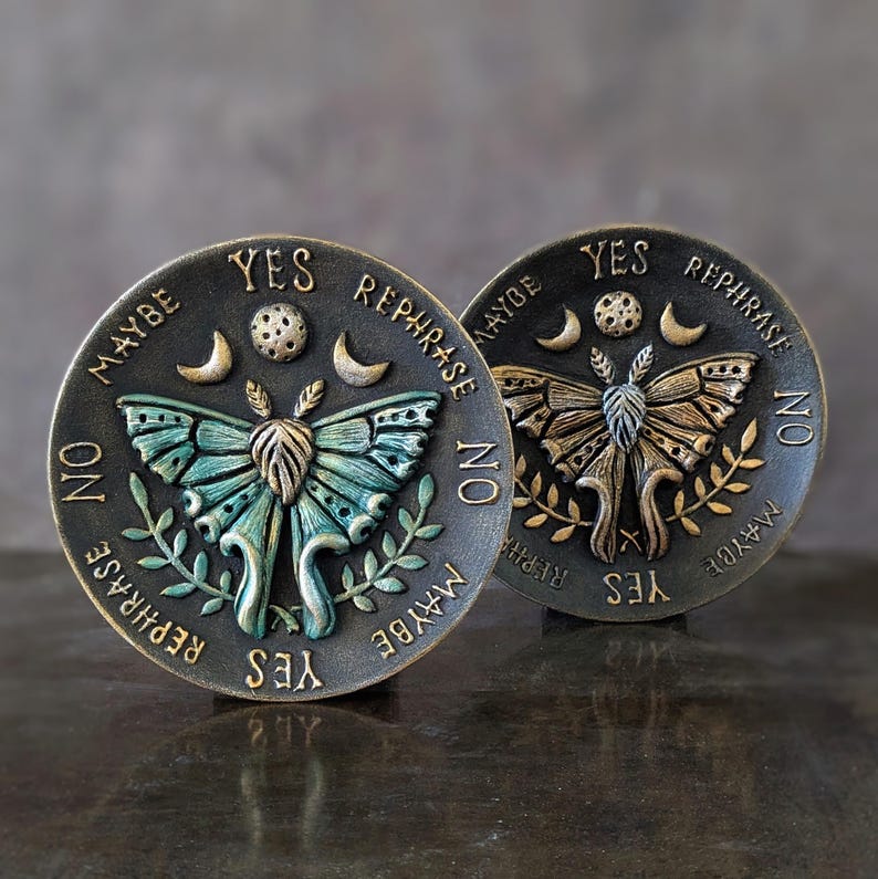 May include: Two circular dark metal plaques featuring a central moth relief, a moon, and stars. The text YES, NO, MAYBE, and REPHRASE is embossed around the perimeter. One moth is teal and gold, while the other is entirely gold-toned.