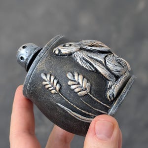 Puede incluir: Un salero decorativo pintado a mano con un relieve detallado de un conejo y espigas de trigo. El salero es principalmente gris oscuro con detalles plateados y dorados. La parte superior es de metal plateado perforado.